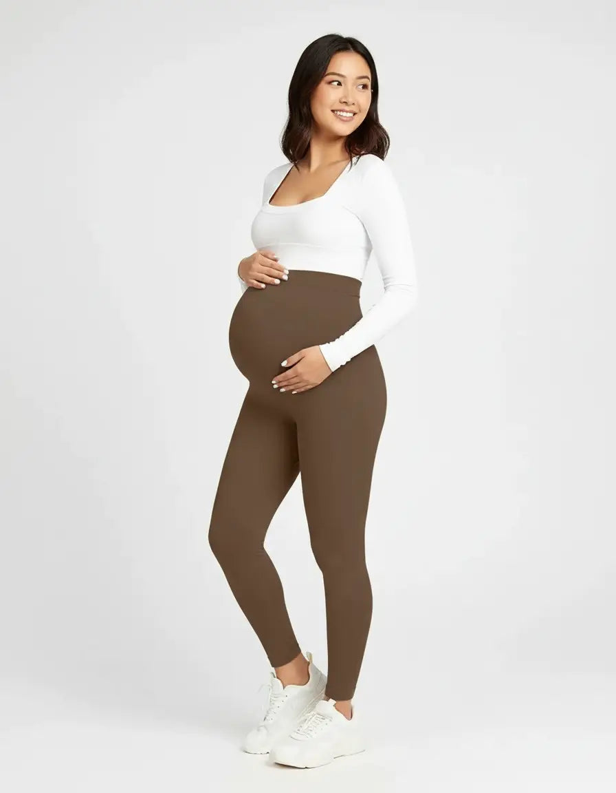 Smiling pregnant woman in soft-stretch, full-length brown maternity leggings and a white long-sleeve top.