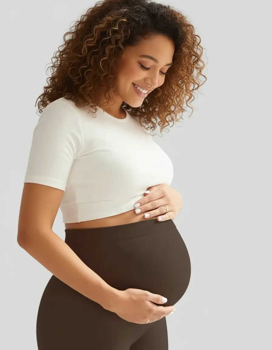 Smiling pregnant woman in a white crop top and dark brown soft-stretch full-length maternity leggings.