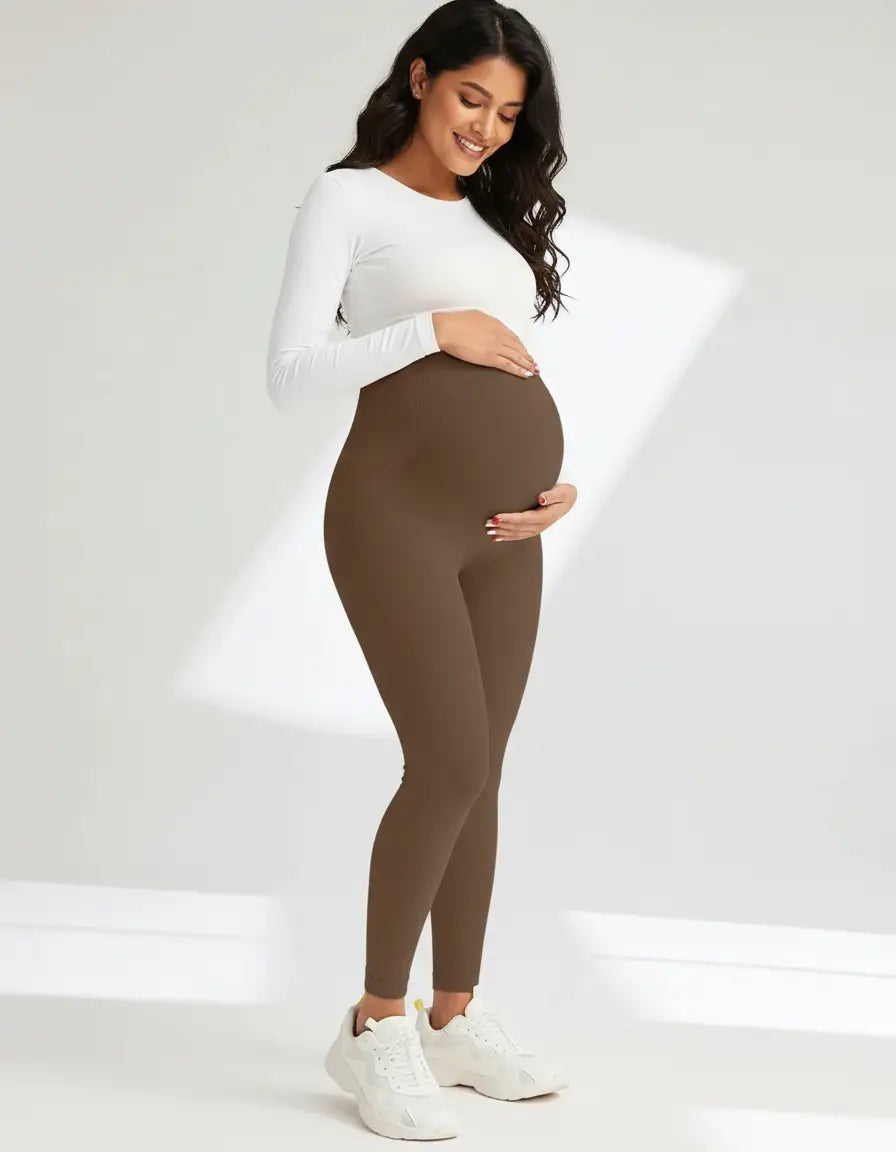 Smiling pregnant woman in soft-stretch, full-length brown maternity leggings and a white long-sleeve top.