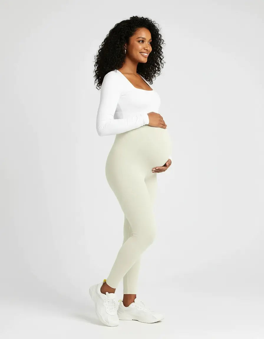 Smiling pregnant woman in soft-stretch, full-length light beige maternity leggings and a white long-sleeve top.