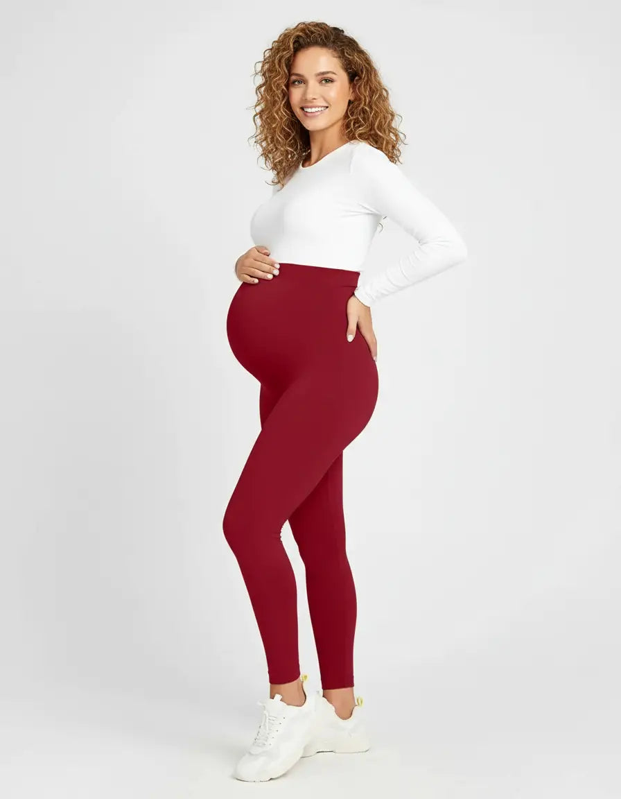 Smiling pregnant woman in a white long-sleeve top and soft-stretch full-length burgundy maternity leggings.