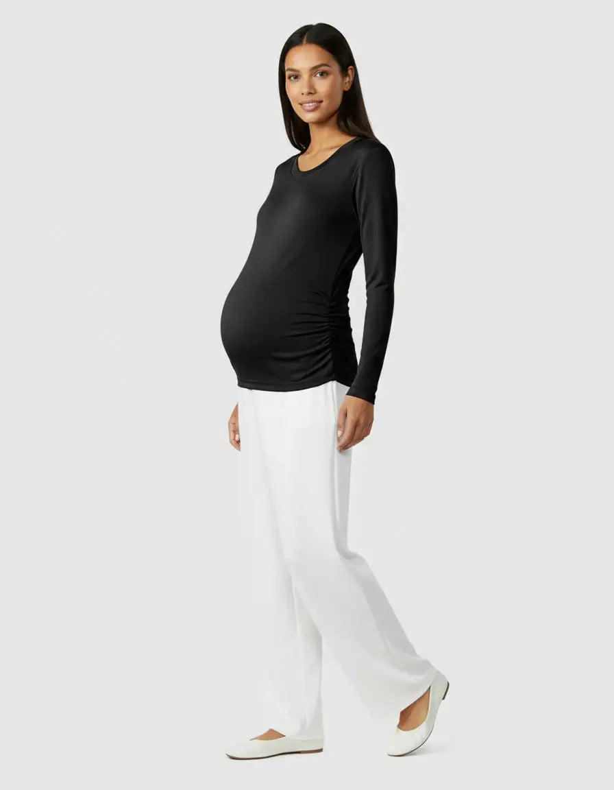 Smiling pregnant woman in a black long-sleeve thermal maternity top with side ruching and white pants.