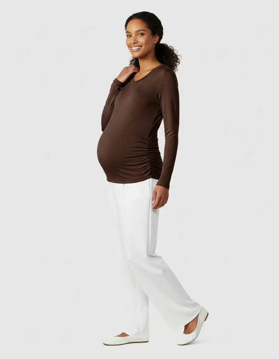 Smiling pregnant woman wearing a brown long-sleeve thermal maternity top with side ruching and white pants.