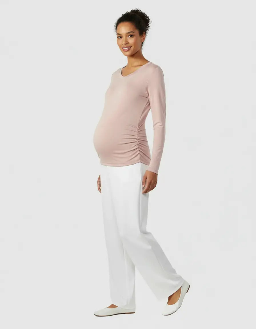Smiling pregnant woman in a soft pink thermal long sleeve maternity top with ruched sides and white pants.