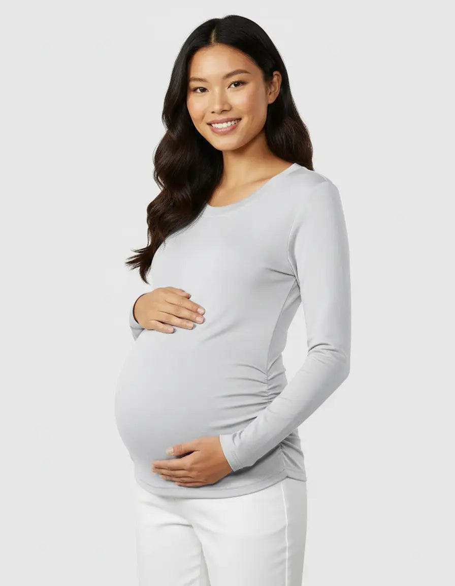 Smiling pregnant woman in a light grey thermal long sleeve maternity top with side ruching and white pants.