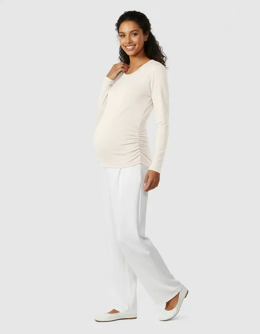Smiling pregnant woman in a cream thermal long-sleeve maternity top with side ruching and white pants.