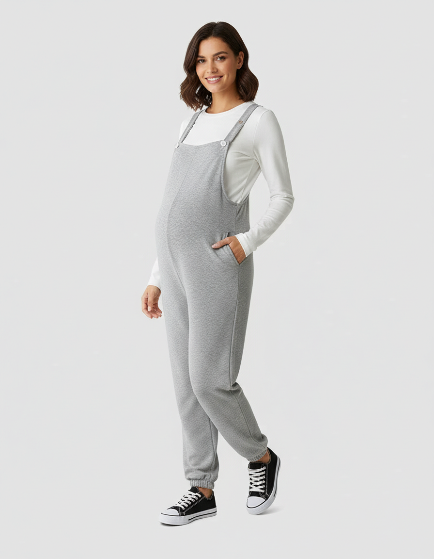 Smiling pregnant woman in gray suspended-strap wide-leg maternity overalls with pockets and a white long-sleeve top.