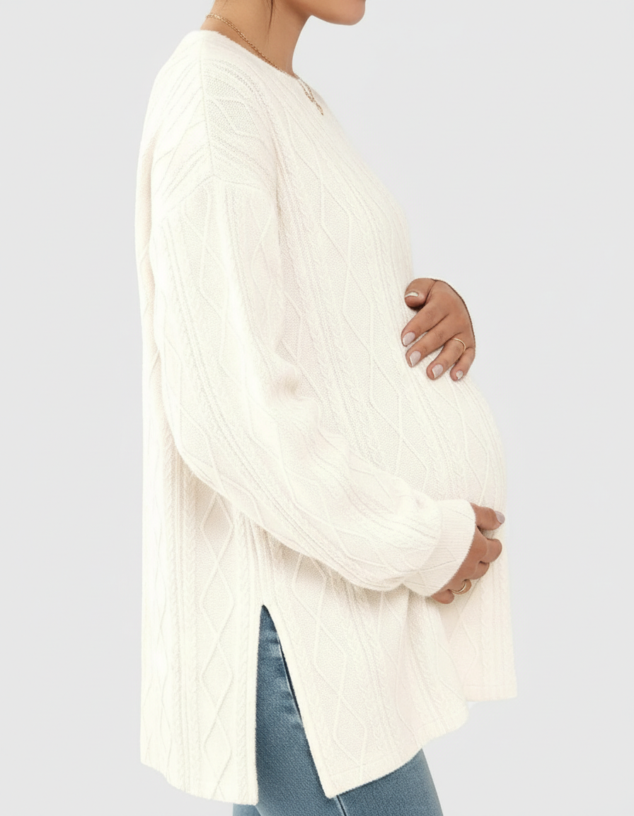 Pregnant woman wearing a cream cable knit loose-fit maternity top with side slits and blue jeans.