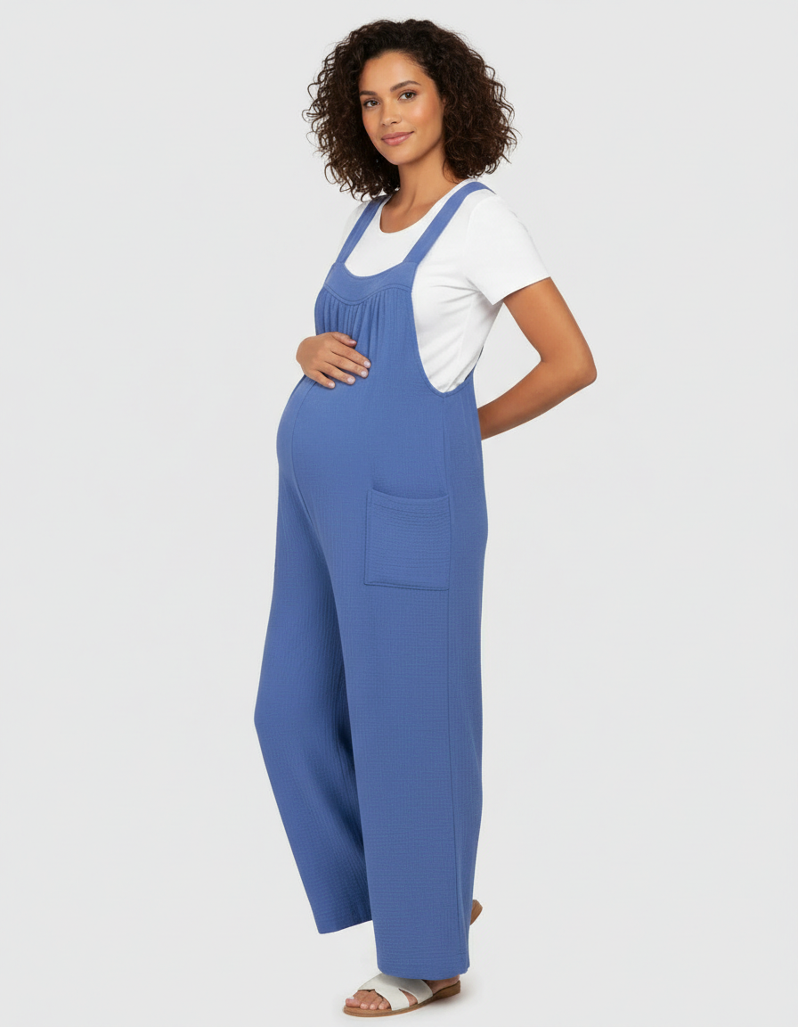 Pregnant woman in a blue textured wide-leg maternity jumpsuit with a white tee, side pockets, and tie-back straps.