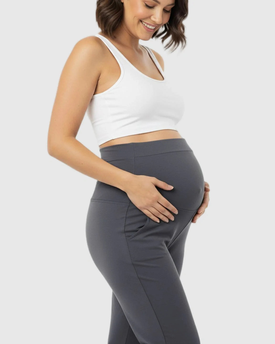 Smiling pregnant woman in a white crop top and charcoal gray stretch flared maternity pants with pockets.