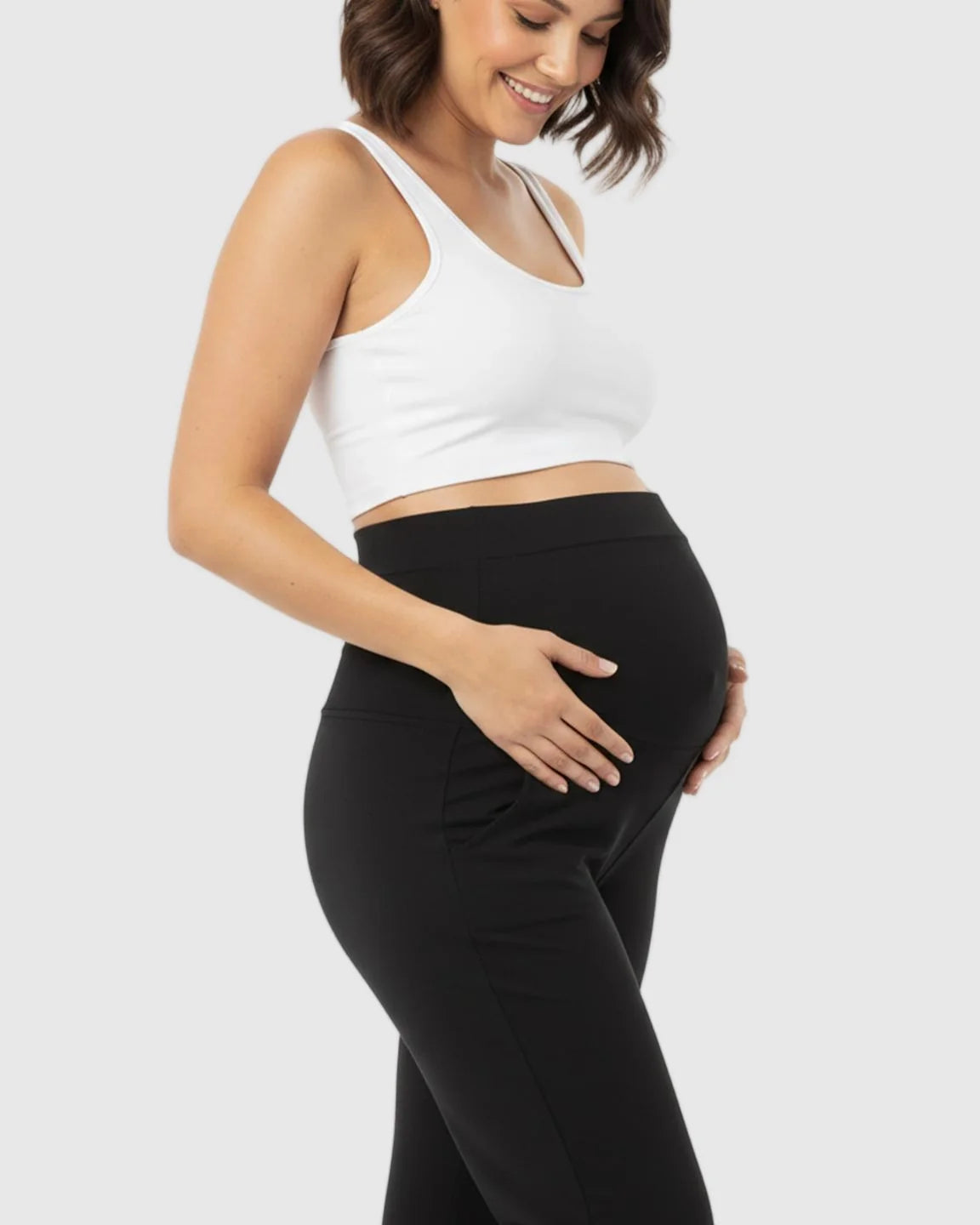 Smiling pregnant woman wearing black stretch flared maternity pants with an over-belly waistband and a white crop top.