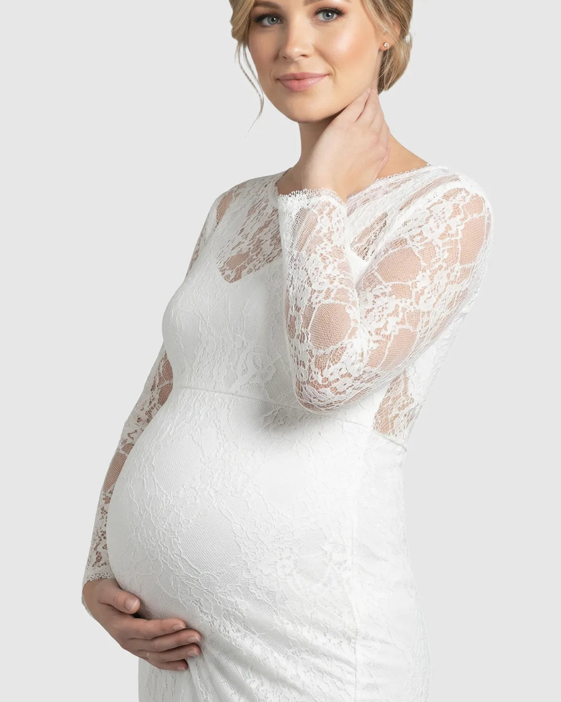 Pregnant woman in a white sheer lace trumpet maternity gown with long sleeves, holding her baby bump.