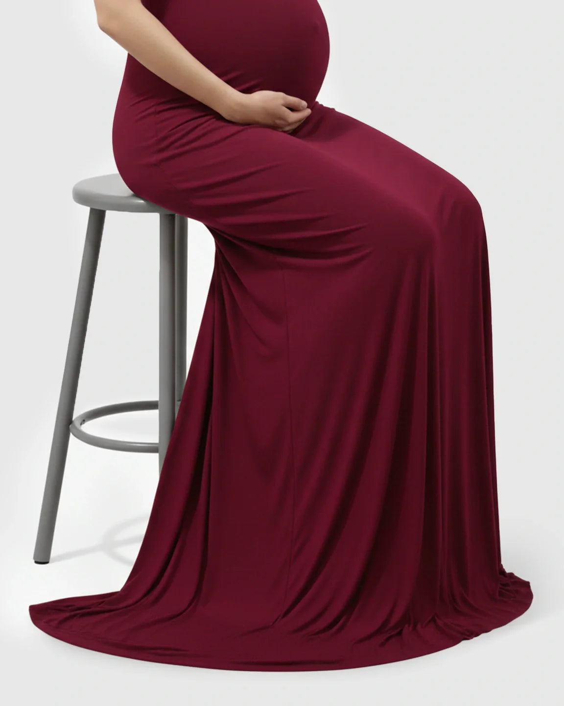 Pregnant woman in a burgundy off-shoulder bodycon maternity mermaid gown, seated on a stool.