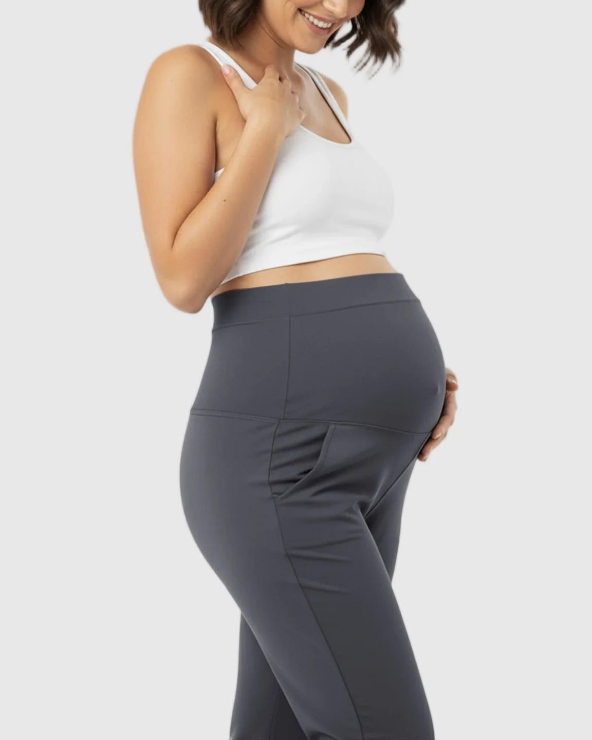 Smiling pregnant woman wearing dark gray stretch flared maternity pants with pockets and a white crop top.