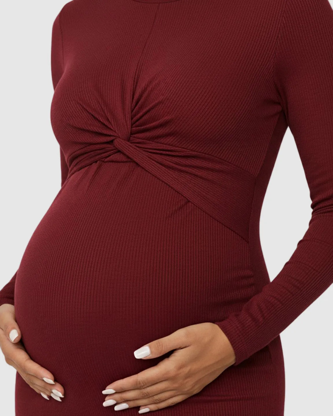 Maternity model in a long-sleeve, burgundy ribbed bodycon dress with a twist-front detail, cradling her bump.