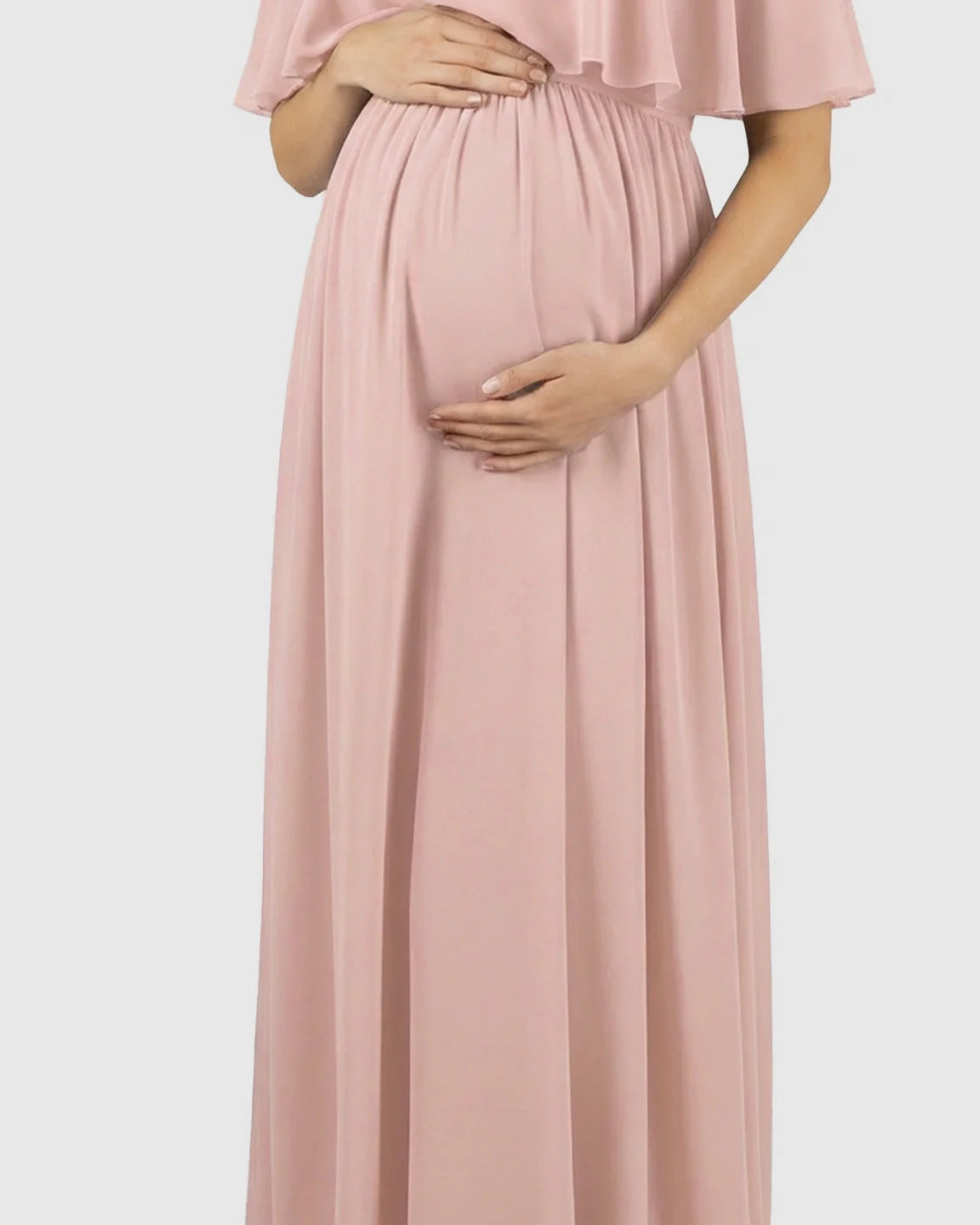 Maternity model in a flowy, blush pink chiffon off-shoulder bohemian dress, cradling her baby bump.