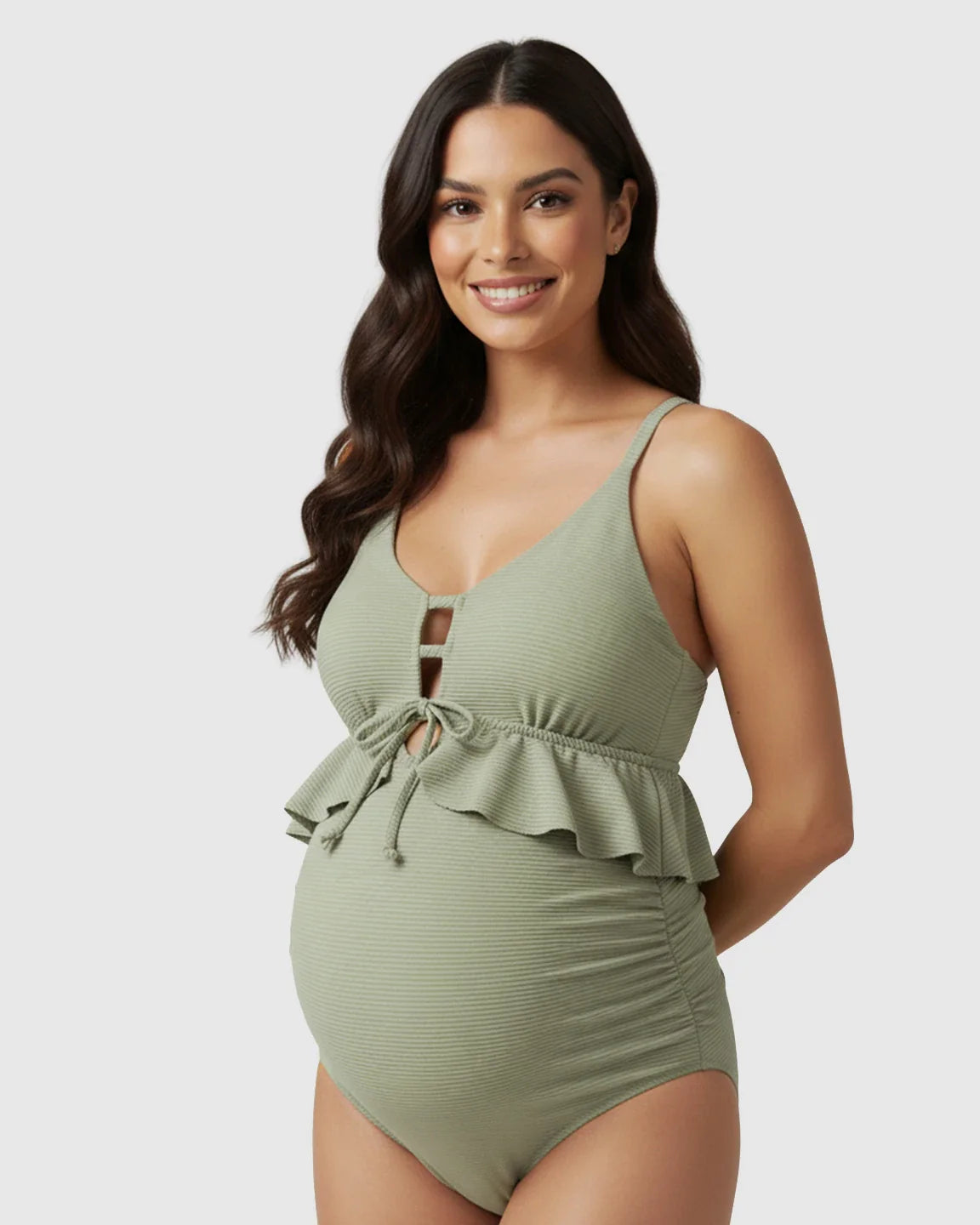 Smiling pregnant woman in a sage green ribbed tankini swimsuit with a plunging V-neck, keyhole tie, and peplum ruffles.