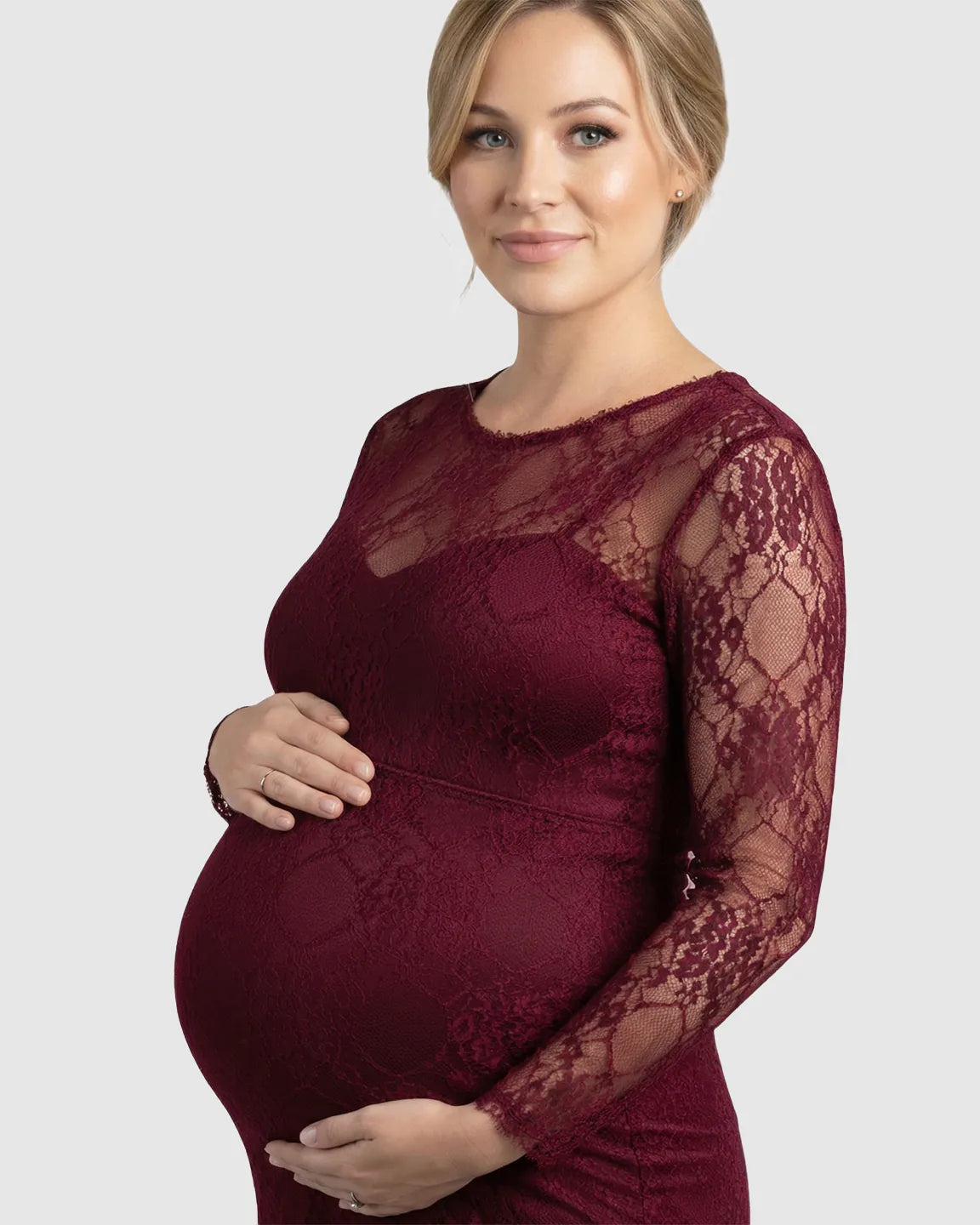 Pregnant woman in a sheer burgundy lace trumpet maternity gown with long sleeves, cradling her bump.
