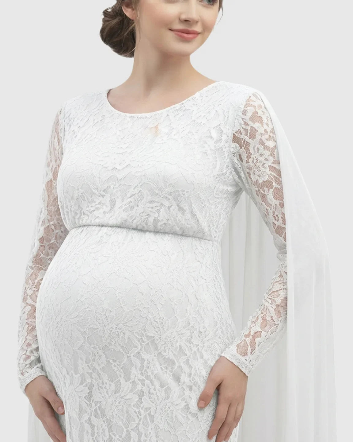 White lace maternity gown with long sleeves, empire waist, and a flowing chiffon cape trailing from the left shoulder.