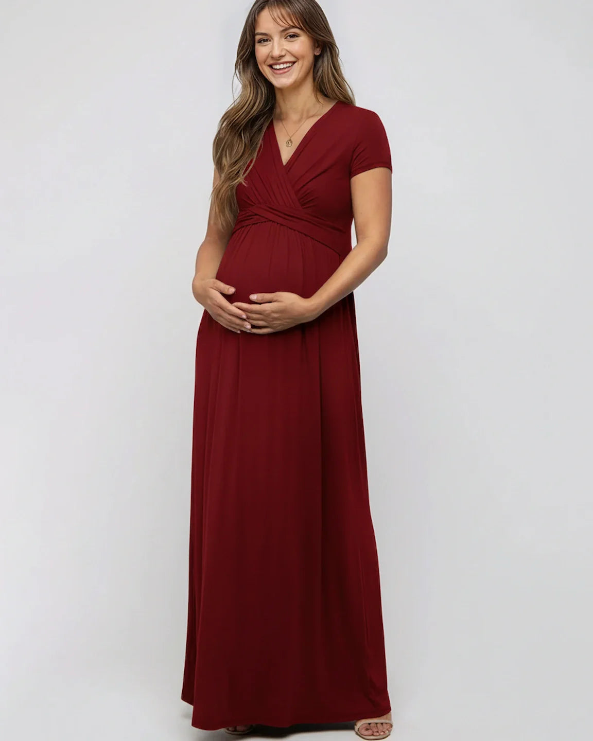 Smiling pregnant woman in a red twist-front draped maternity nursing maxi dress, holding her bump.