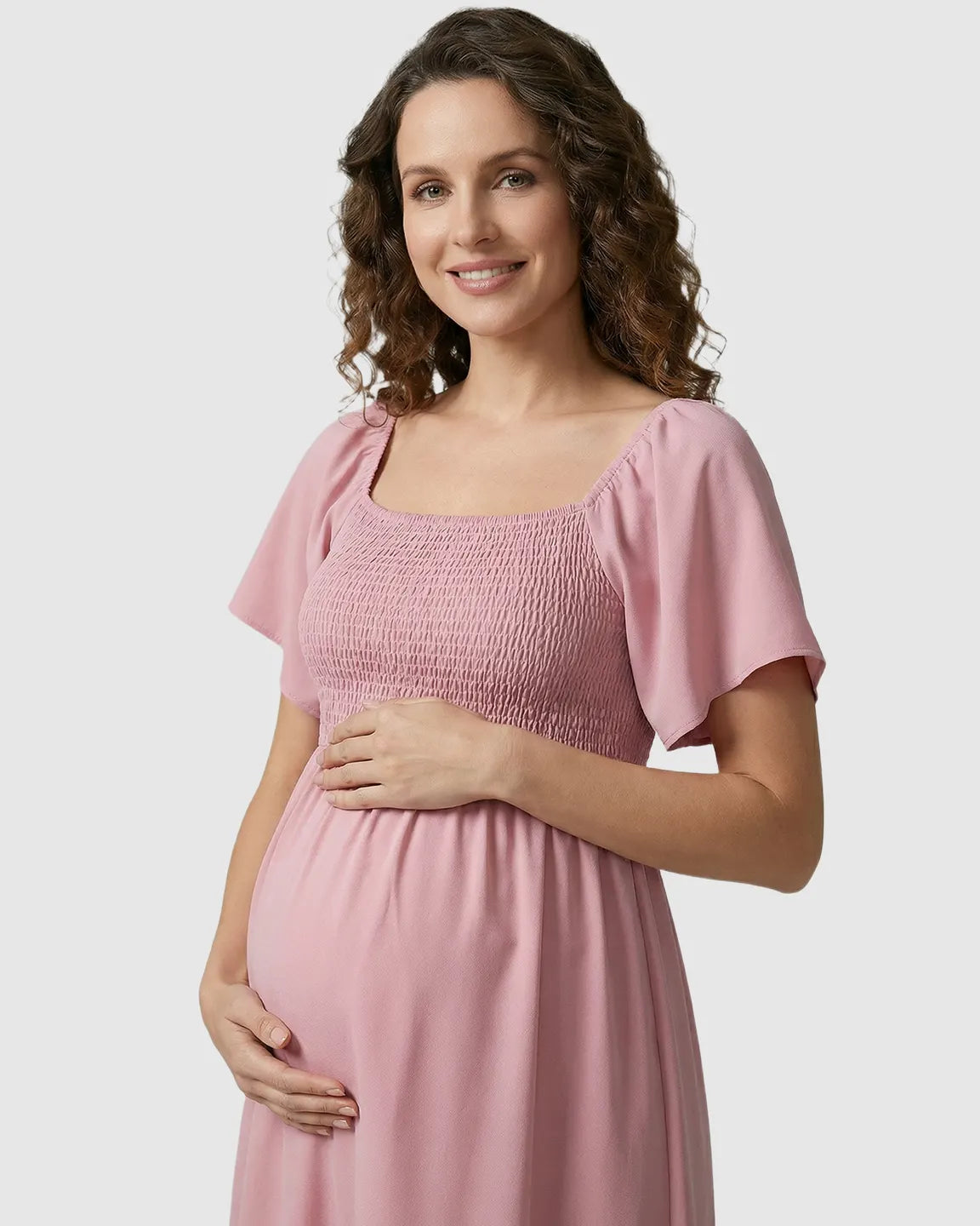 Smiling pregnant woman in a pink flutter-sleeve smocked maternity midi dress, holding her belly.