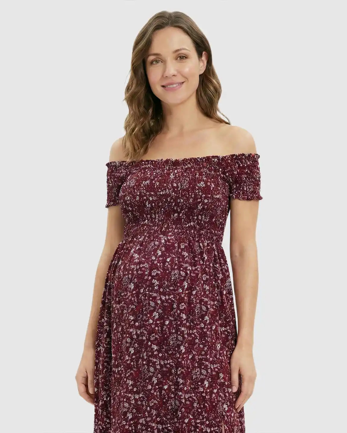 Smiling pregnant woman in a red floral off-shoulder maternity maxi dress with a side slit.