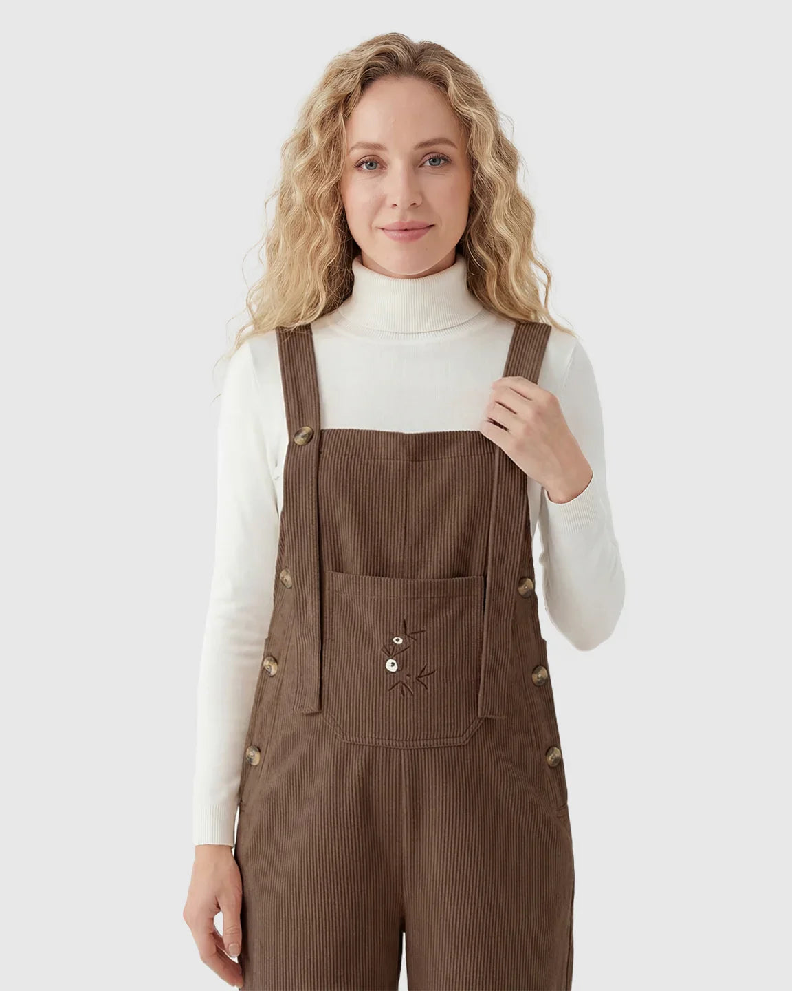 Woman in brown corduroy maternity overalls with adjustable straps and front pocket, over a white turtleneck.
