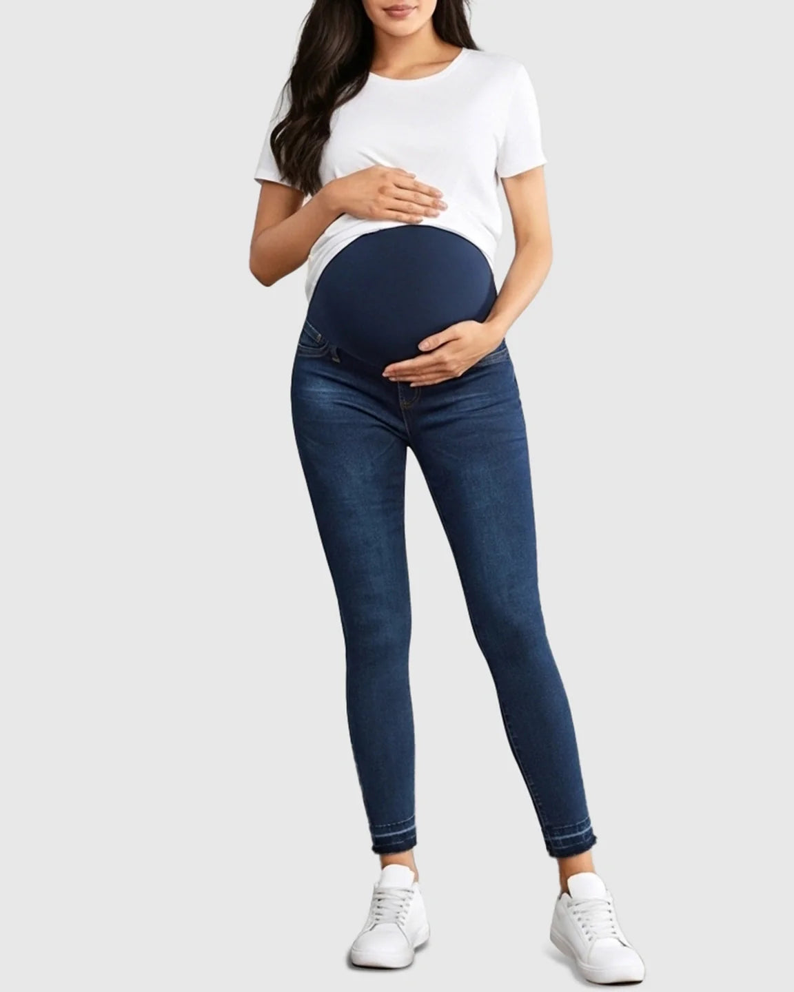 Pregnant woman wearing dark wash skinny maternity jeans with a navy high-waist elastic panel and frayed hems.