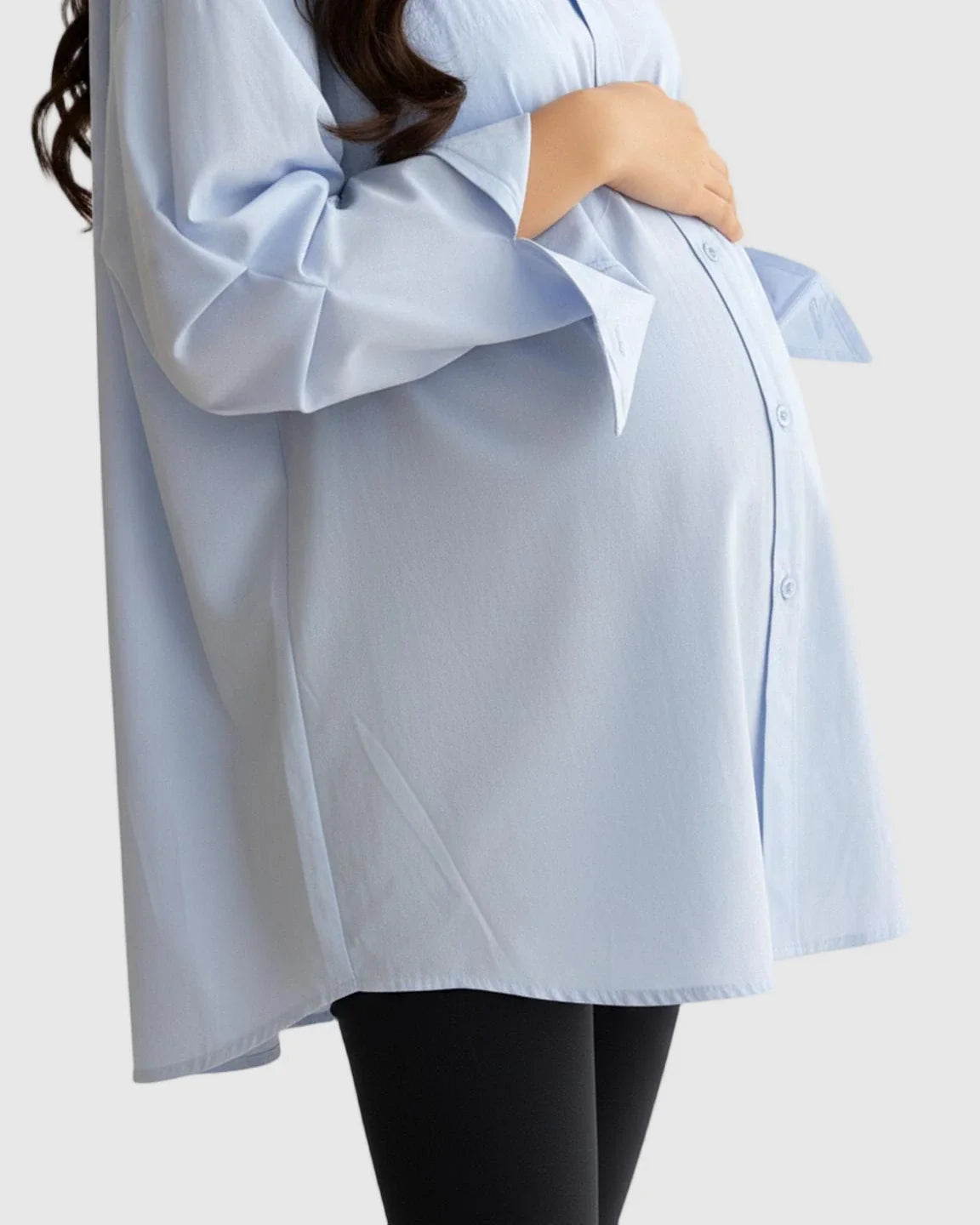 Maternity model in a light blue cotton lapel-collar button-front tunic over black leggings, cradling her bump.