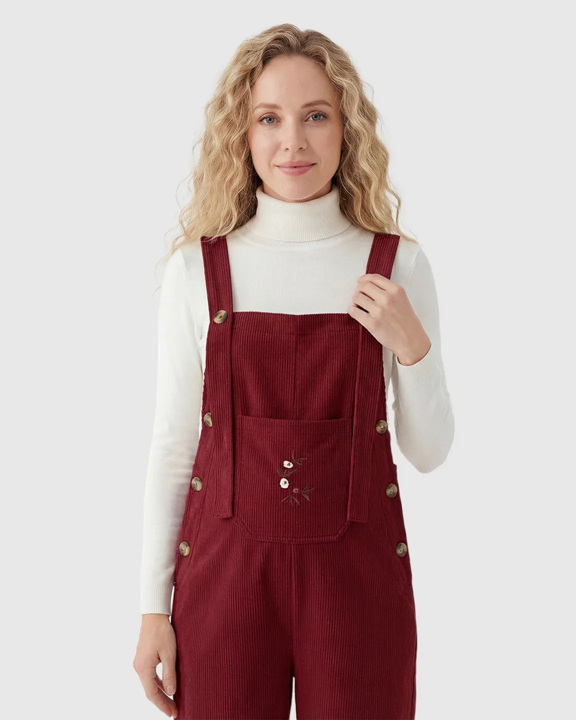 Woman in red corduroy maternity overalls with adjustable straps, side buttons, and embroidered pocket, over a white turtleneck.
