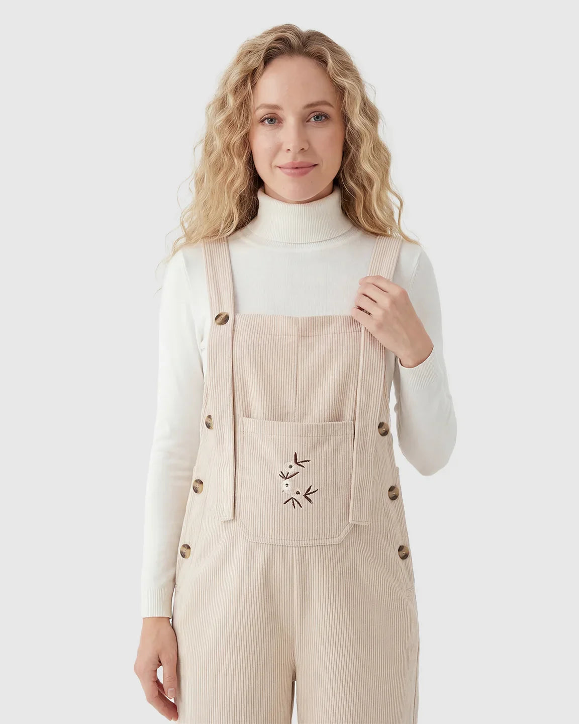 Blonde woman in beige corduroy maternity overalls with adjustable straps, brown buttons, and floral embroidery.