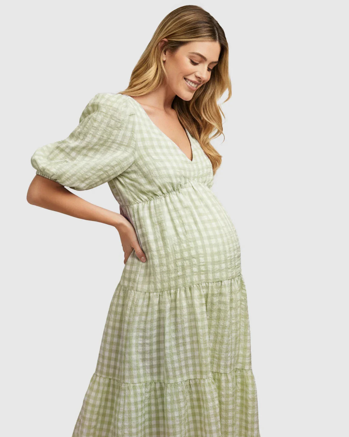Smiling pregnant woman in a green and white checkered maternity maxi dress with puffed sleeves and a V-neck.