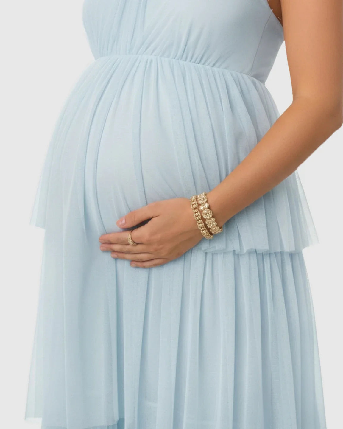Maternity model in a light blue halter neck tiered mesh gown, cradling her bump.