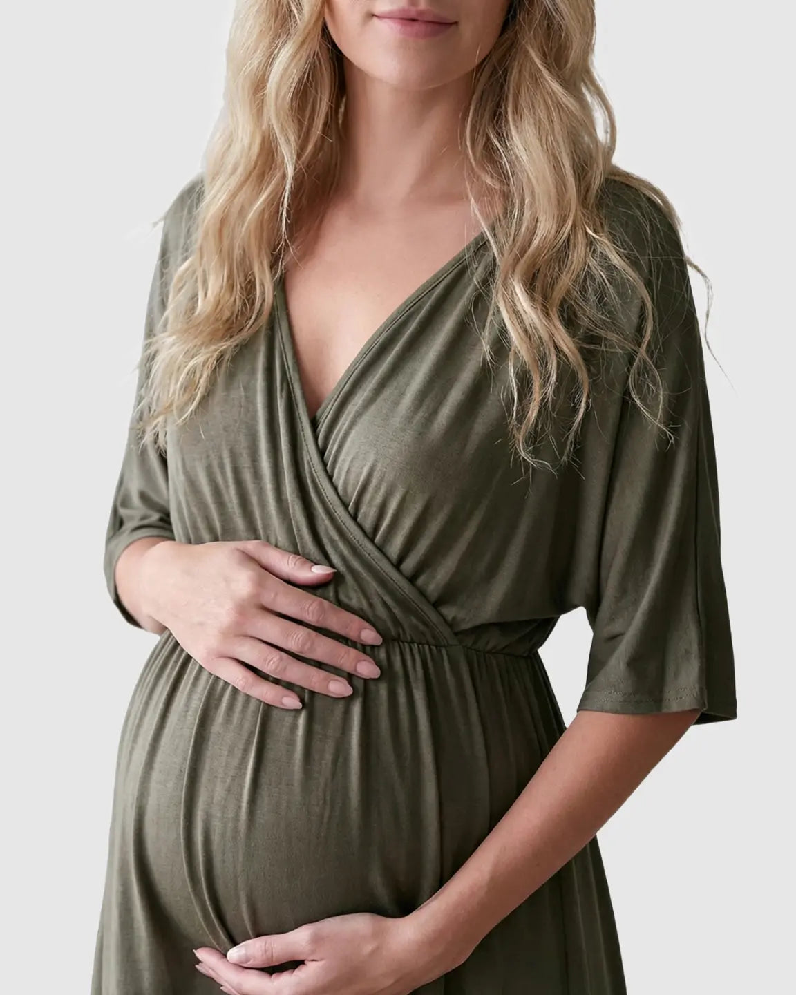 Pregnant woman in a soft olive green cross-front maternity midi dress with 3/4 sleeves, holding her bump.