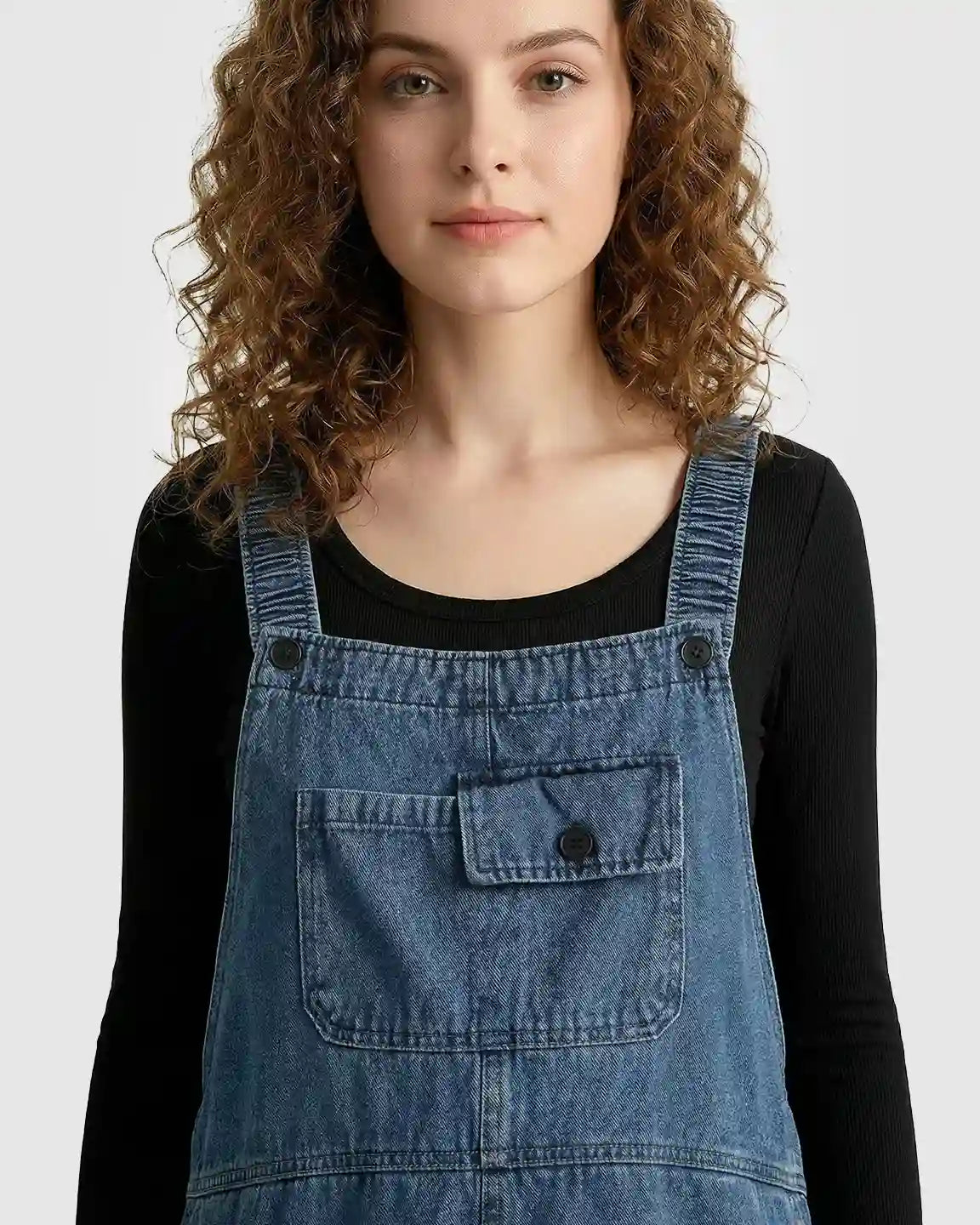 Woman wearing loose-fit blue denim maternity overalls with adjustable straps and front pockets over a black long-sleeve shirt.