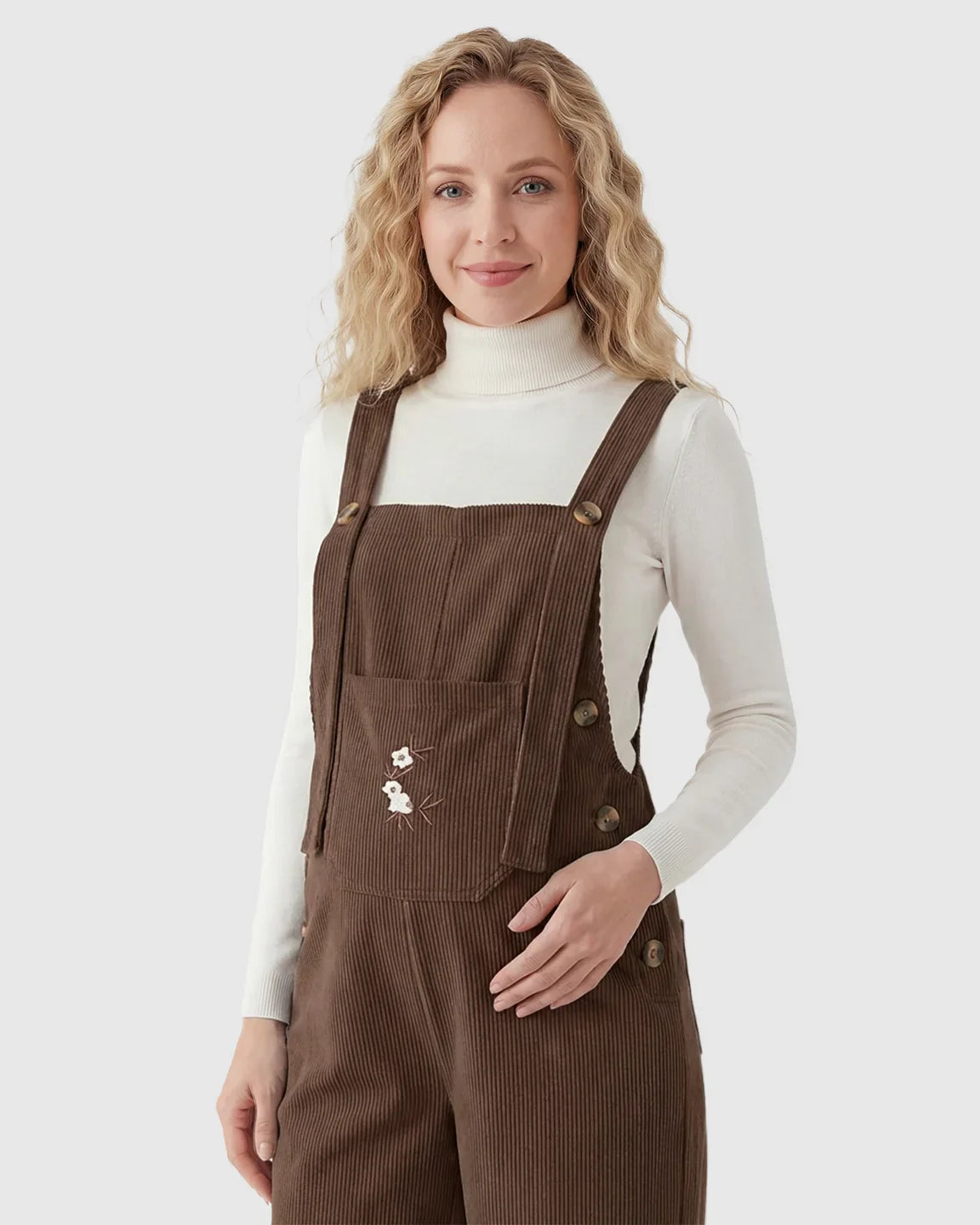Woman in brown corduroy maternity overalls with adjustable straps, white turtleneck, and embroidered flowers.