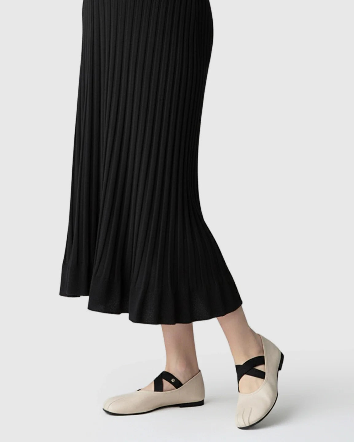 Lower half of a woman wearing a black ribbed knit midi dress and beige flats with black straps.