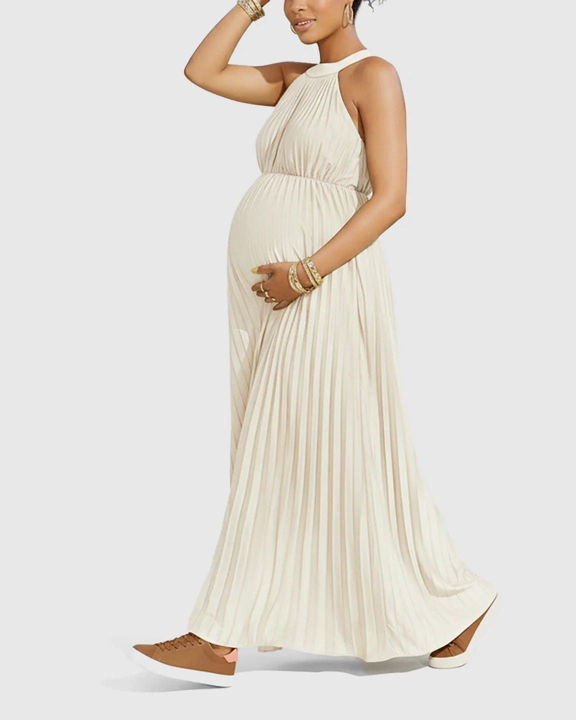 Pregnant model in a cream halter neck pleated maternity maxi dress, made from faux silk, with brown sneakers.