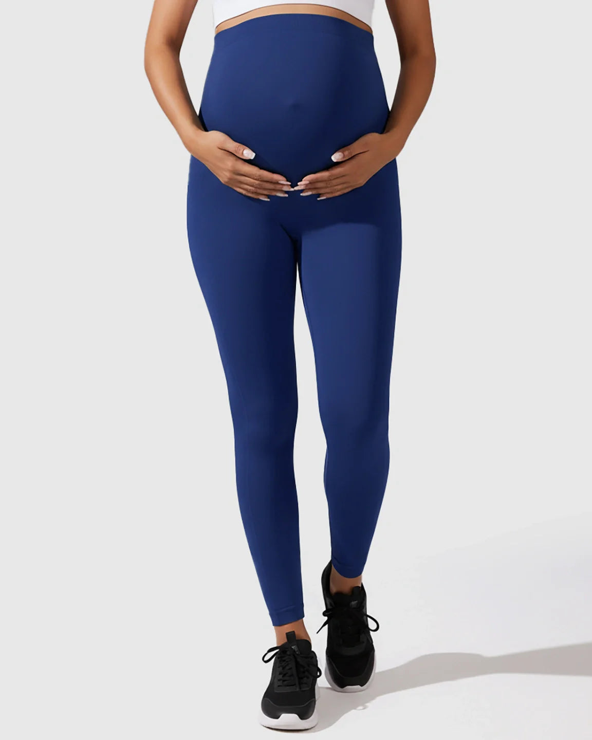 Pregnant woman wearing navy blue seamless, high-waisted maternity leggings, gently holding her bump.