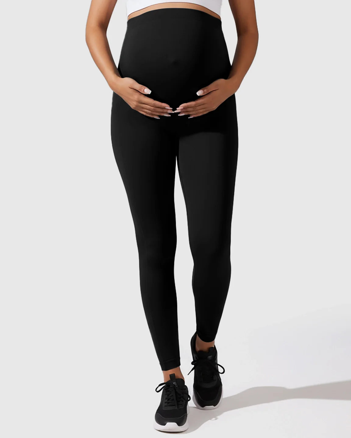 Pregnant woman in black seamless M-shape maternity leggings, gently holding her bump.