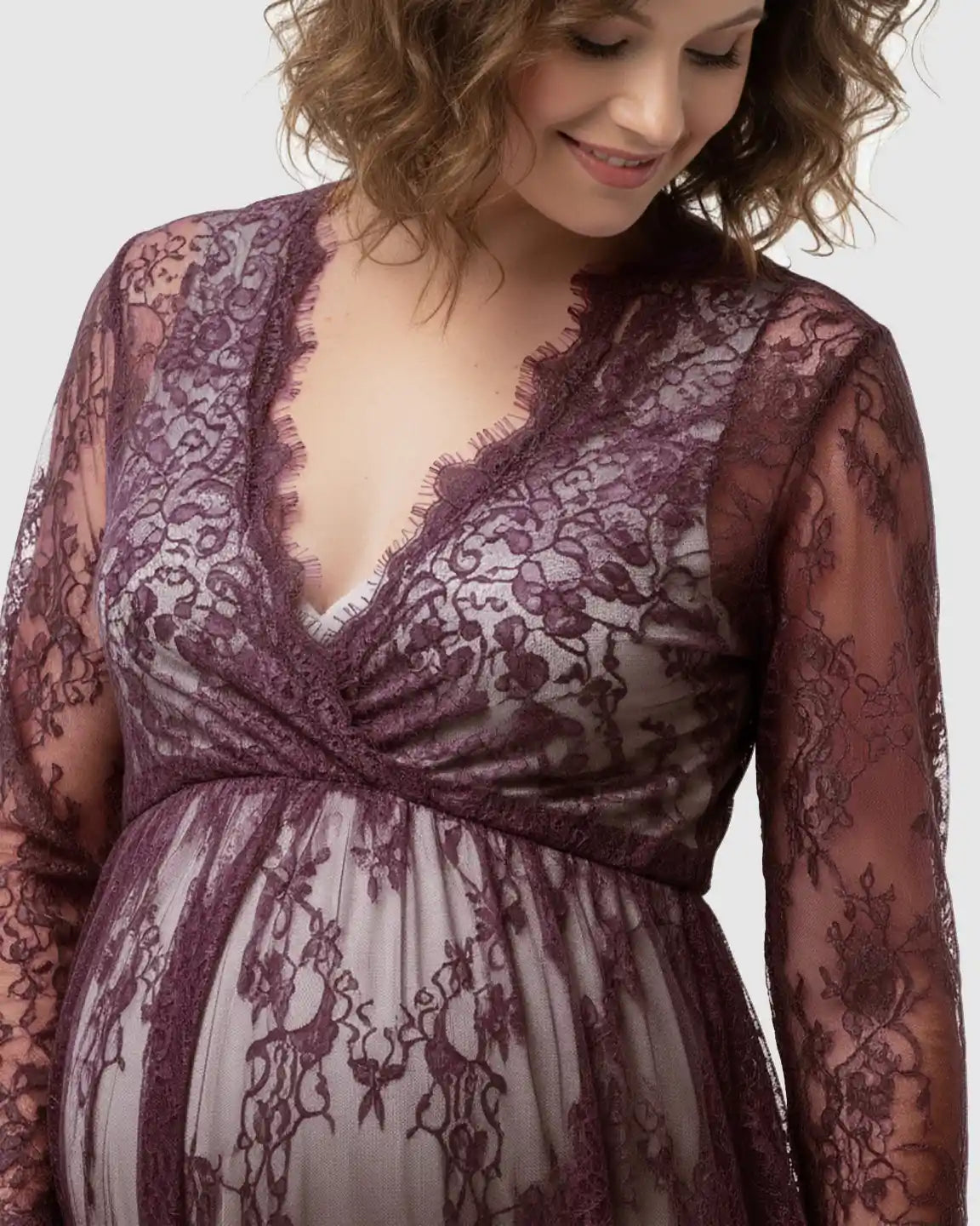 Maternity maxi dress in sheer floral plum lace with an empire waist and V-neck, worn by a smiling pregnant woman.