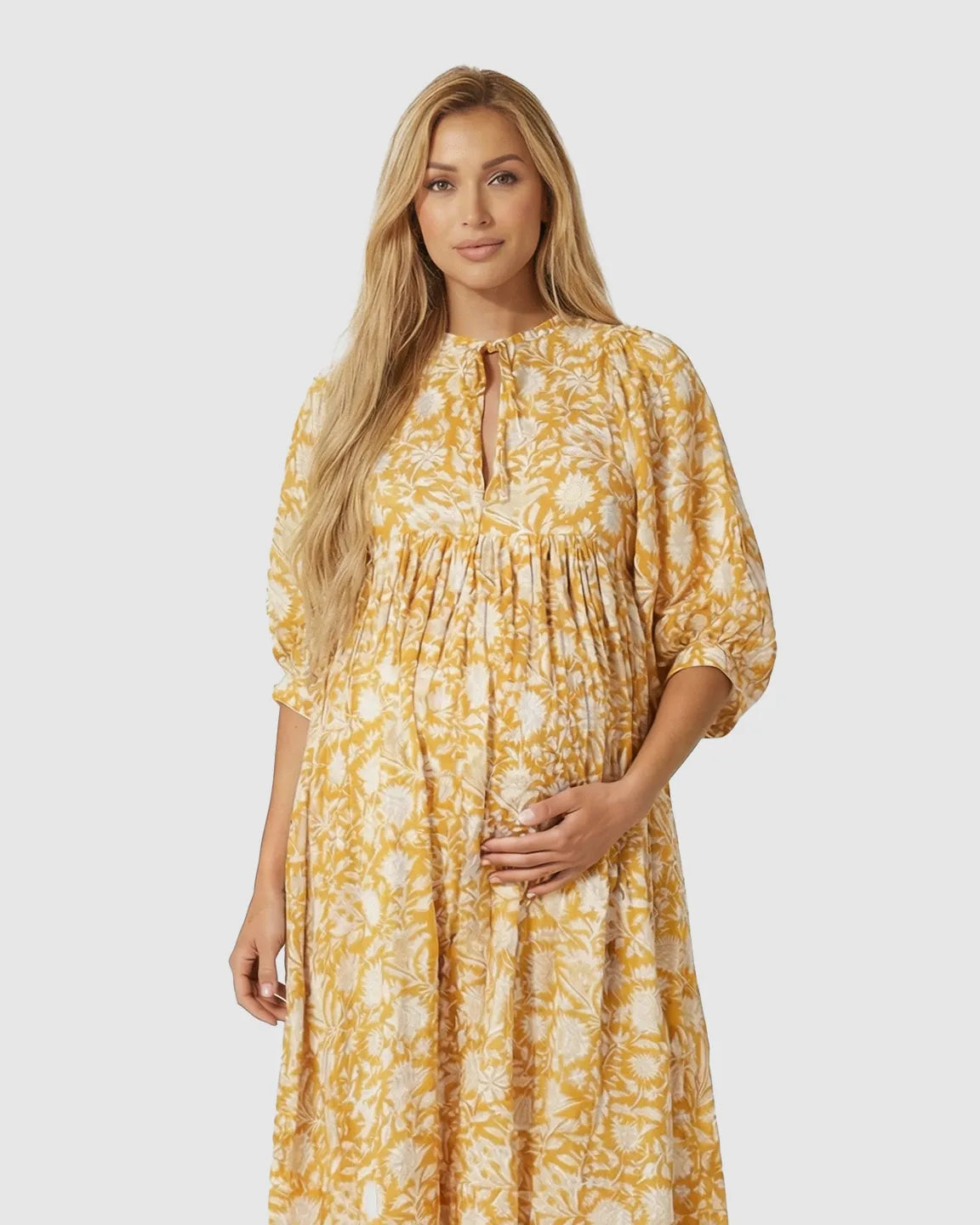 Blonde pregnant woman in a yellow and white floral loose-fit maternity midi dress with gathered shoulders.