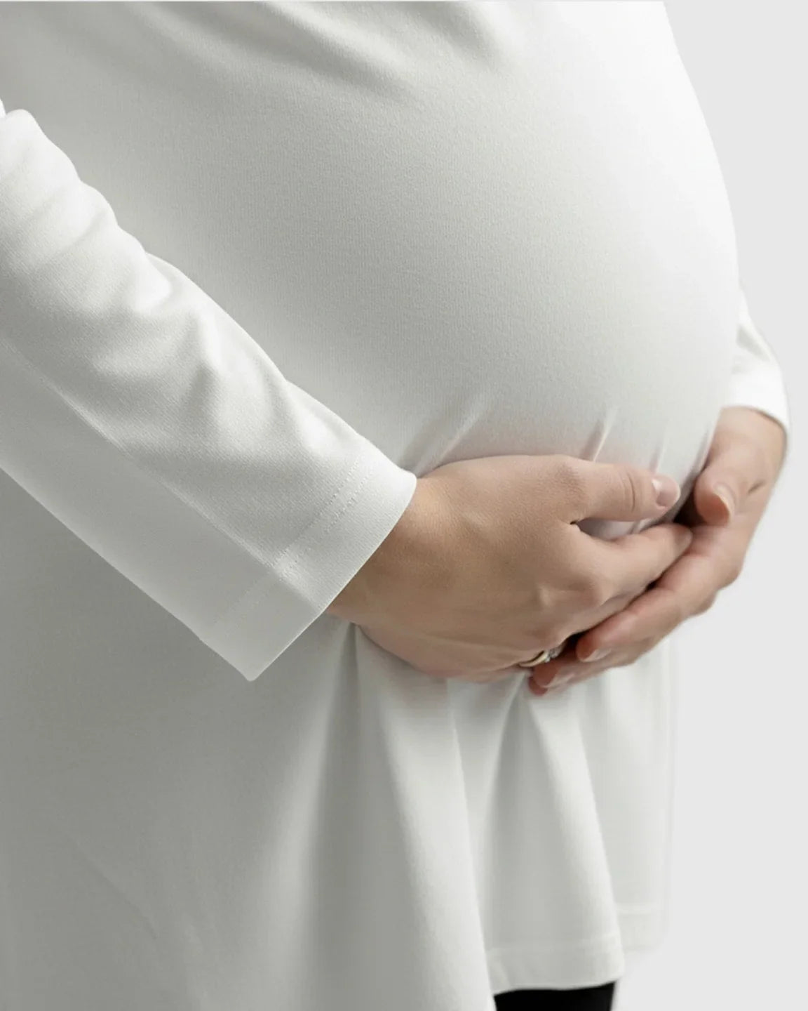 Pregnant woman in a white long-sleeve soft cotton maternity top, hands cradling her bump.