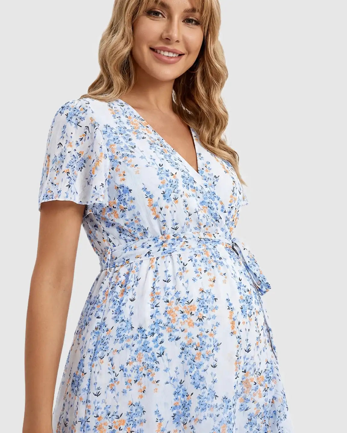 Smiling pregnant woman in a white floral V-neck maternity midi dress with blue and orange flowers, tie waist, and short sleeves.