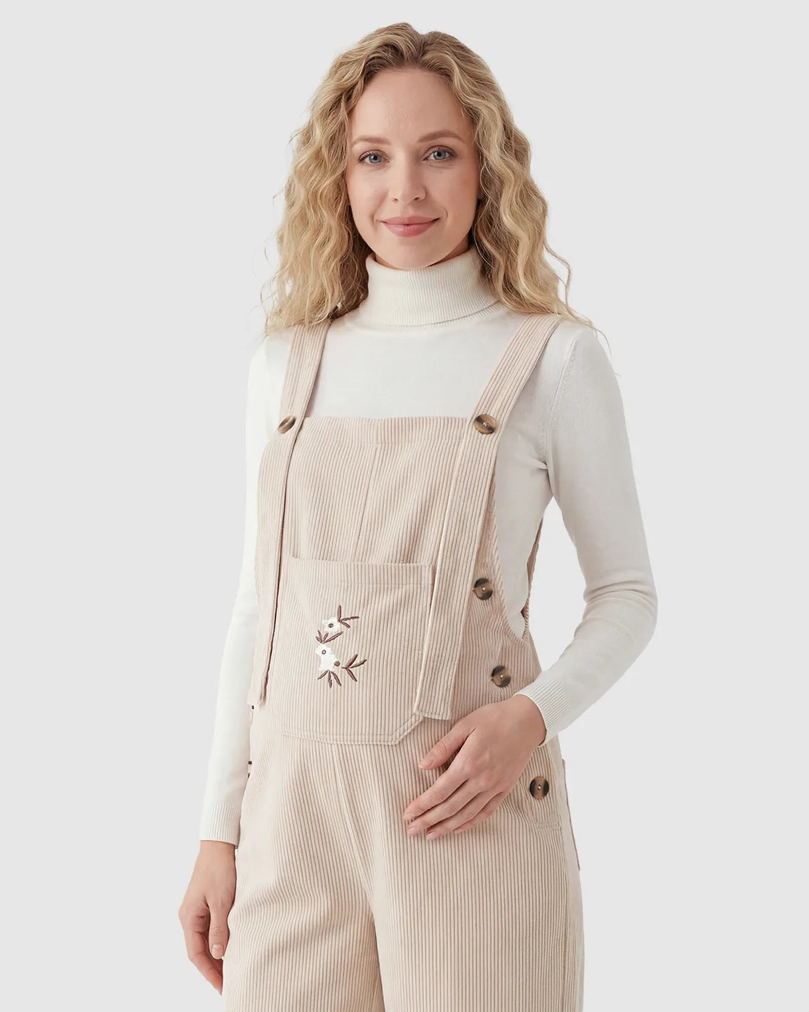 Blonde woman in cream corduroy maternity overalls with adjustable straps, front pocket, and floral embroidery.