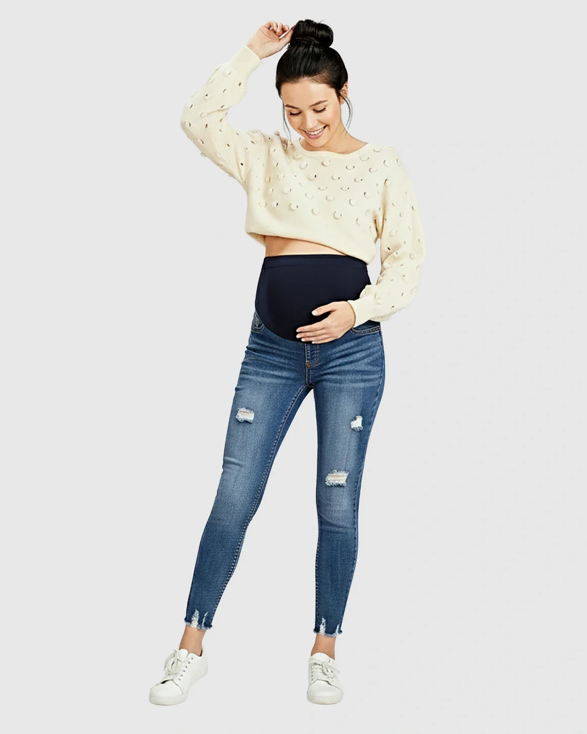 Smiling pregnant woman in distressed skinny maternity jeans with frayed hems and a high-rise elastic waistband.