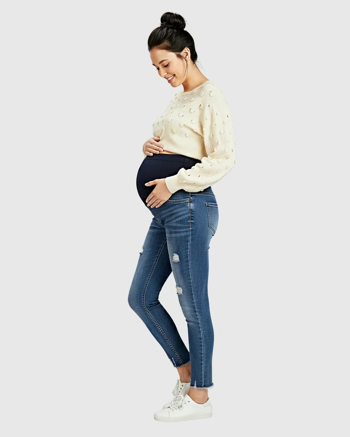 Pregnant woman in blue distressed skinny maternity jeans with frayed hems and a cream sweater.