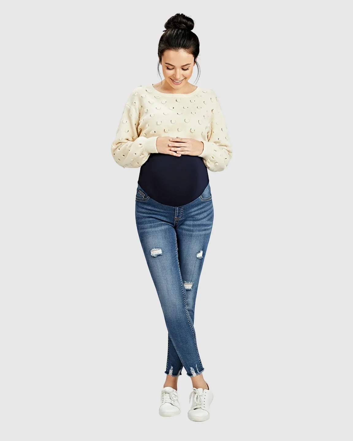 Smiling pregnant woman in distressed skinny maternity jeans with a frayed hem and a cream sweater.