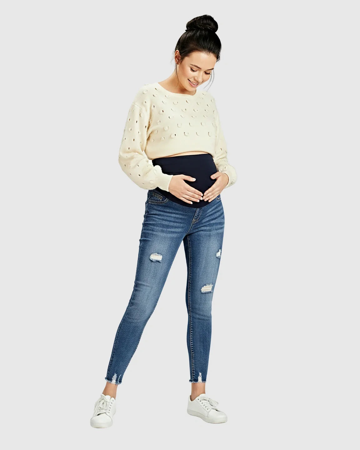 Woman in Bump Society distressed skinny maternity jeans with frayed hems and a cream sweater.