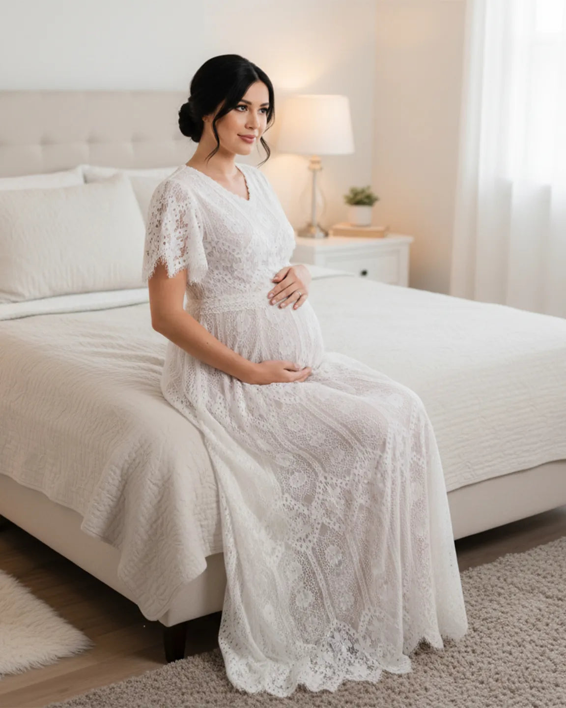 Pregnant woman in a white vintage lace flutter-sleeve maternity gown, sitting on a bed.